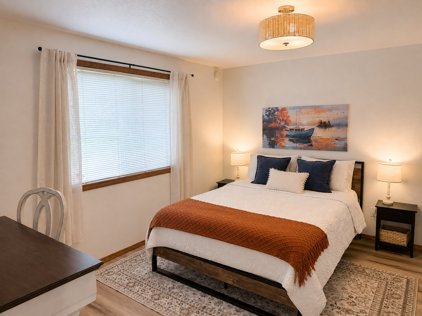 Cozy bedroom with a white bed, navy pillows, rust-colored blanket, window with curtains, bedside lamps, and boat artwork