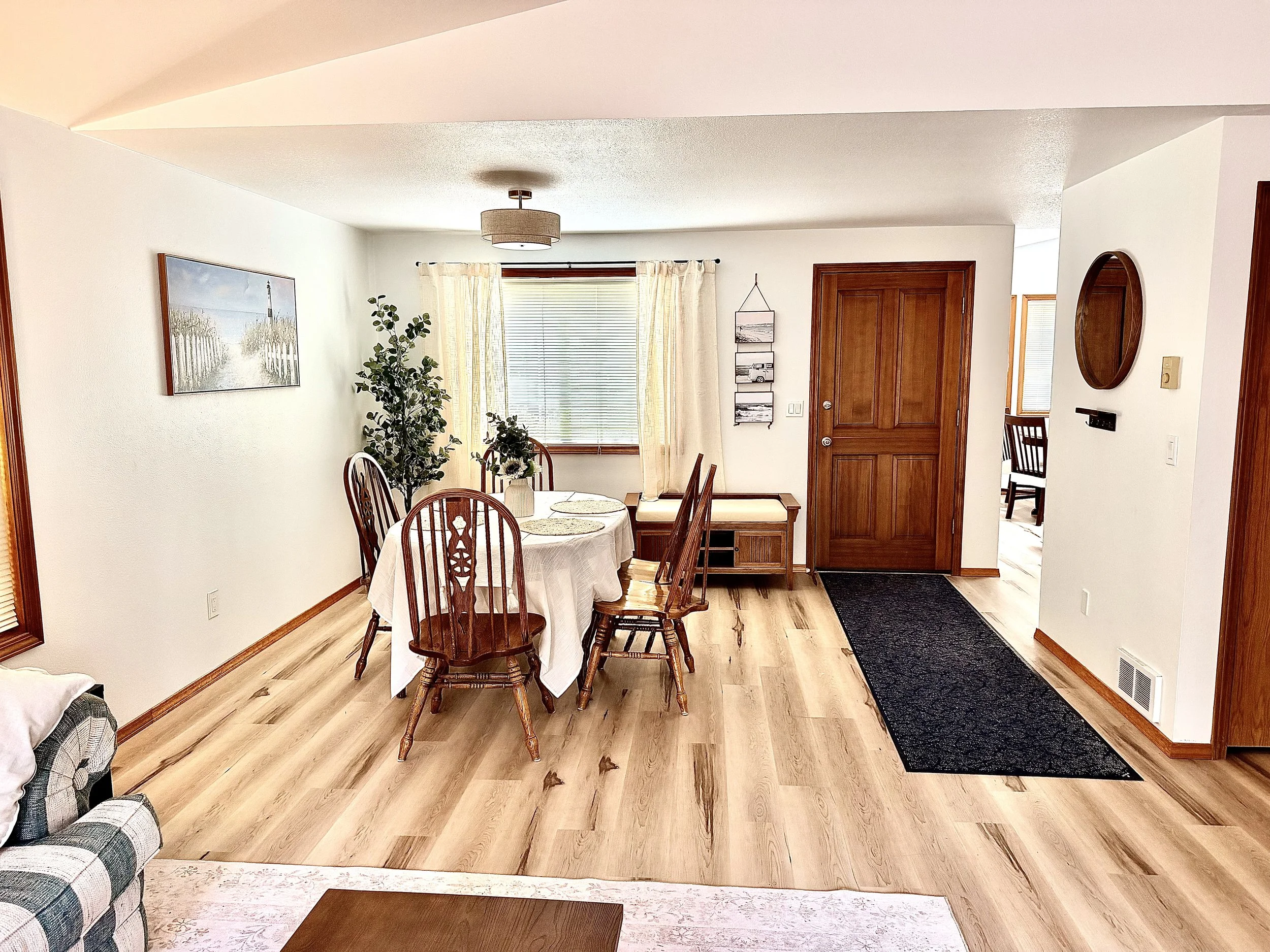 Bright open-plan dining and living room with a wood dining table, rocking chairs, large window, hardwood floors, and entryway.
