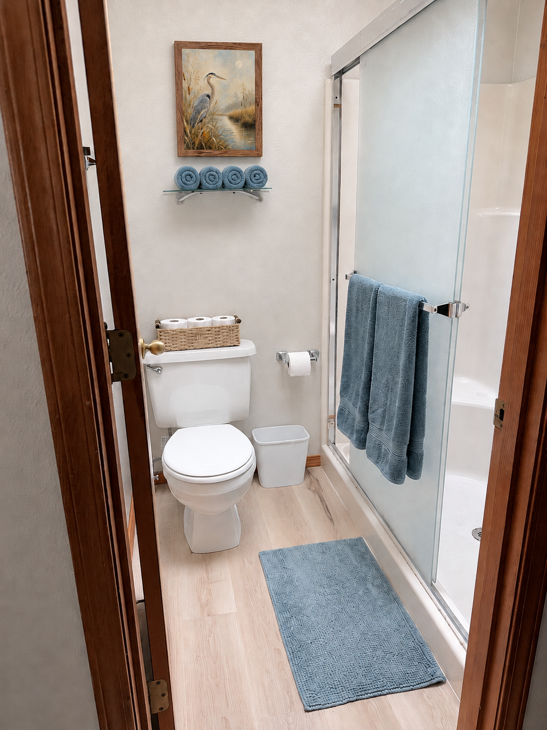 Small bathroom with a white toilet, glass-enclosed shower, blue towels and bath mat, toilet paper basket, and framed heron artwork