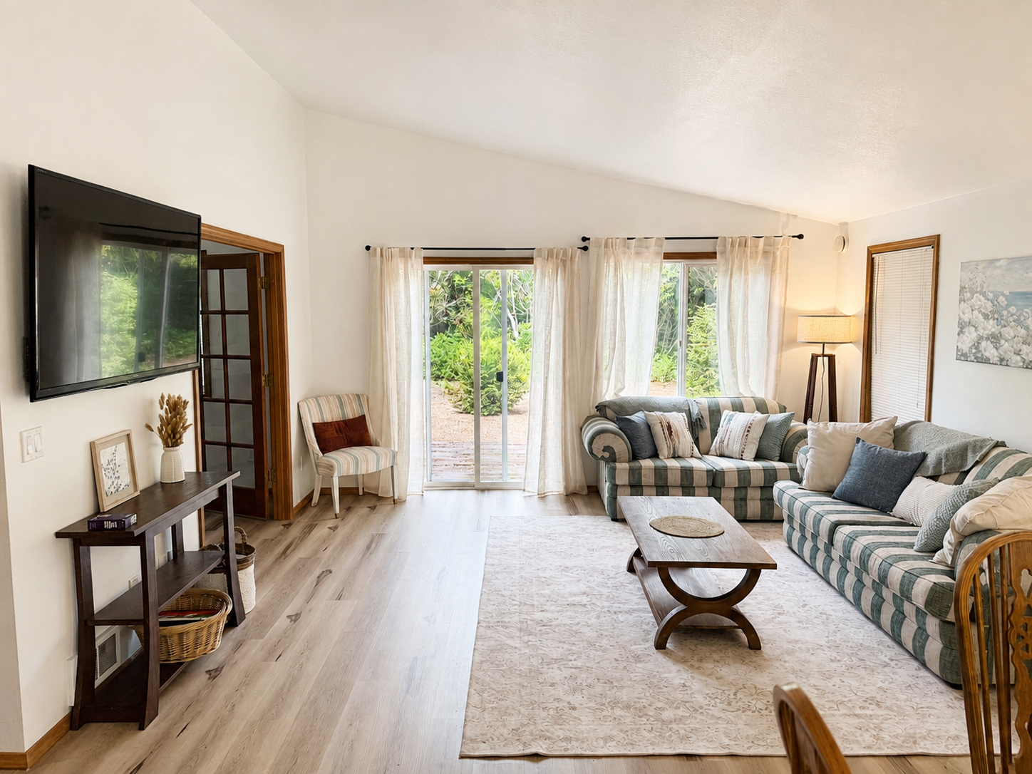 Bright living room with striped sofas, wall-mounted TV, coffee table, area rug, and garden-view sliding glass doors