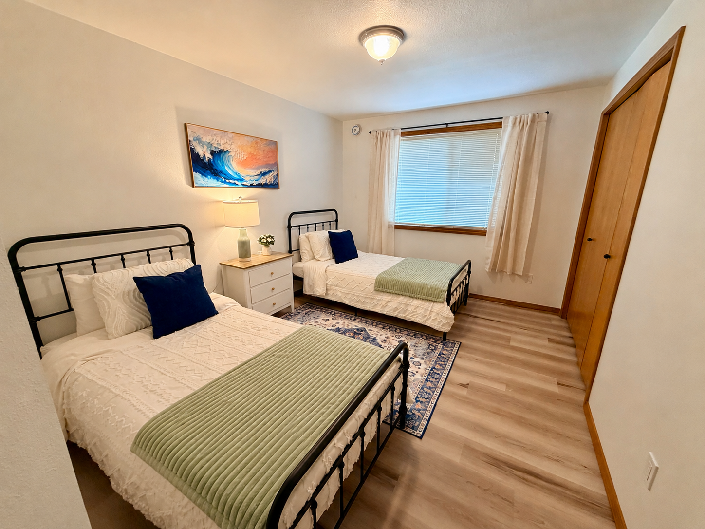Bright twin bedroom with black metal bed frames, white bedding, green blankets, blue accent pillows, wood floors, window, dresser, and ocean-wave artwork