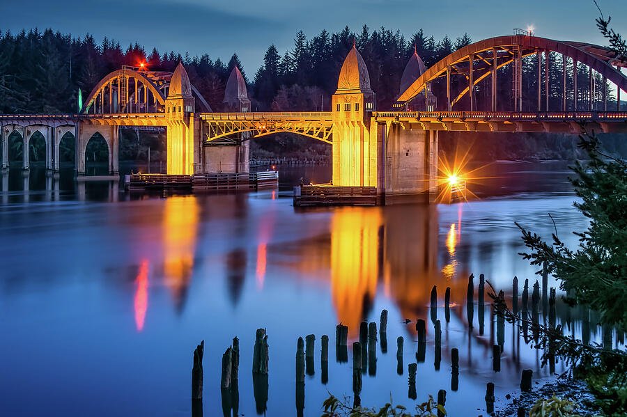 Historic arched bridge illuminated at dusk over calm water, reflecting colorful lights beneath a forested shoreline.