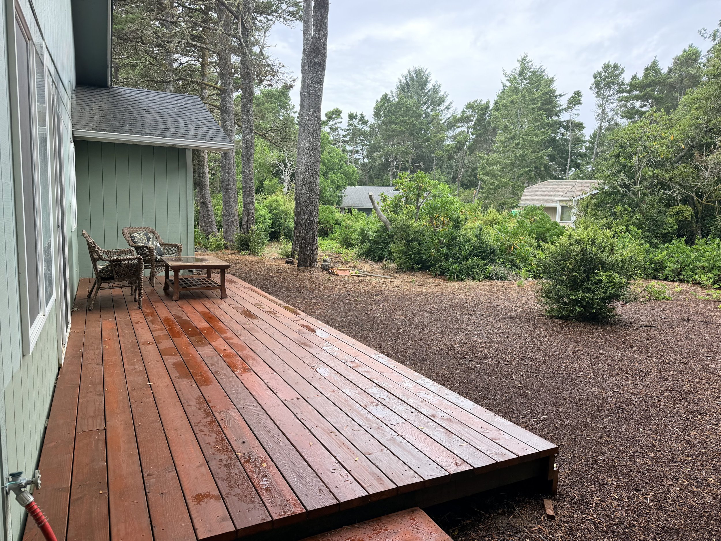 Rainy wooded backyard with a redwood deck, wicker chairs, coffee table, garden beds, and neighboring cottages