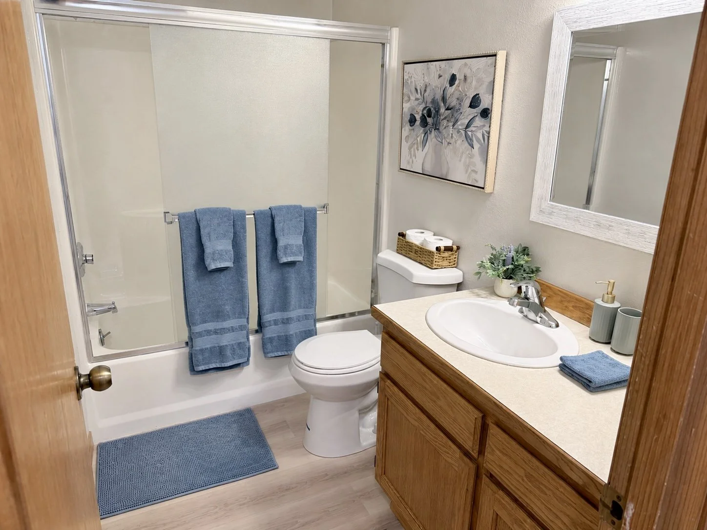 Clean bathroom with bathtub and glass doors, blue towels and bath mat, toilet, wooden vanity, sink, mirror, and decorative plant