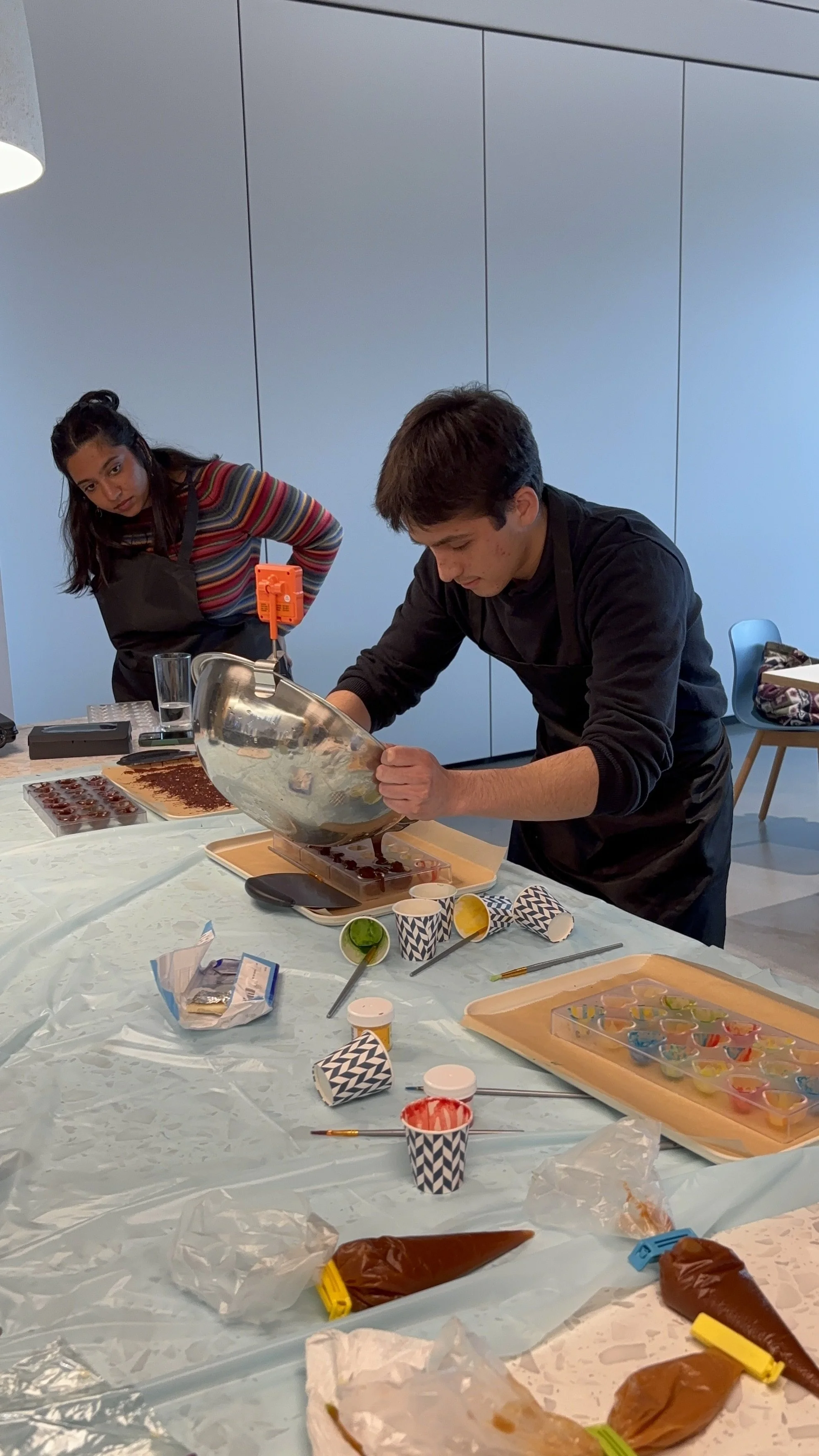 Two people decorating chocolates at a table. One person is pouring chocolate into molds, and the other is standing nearby watching. The table is covered with a plastic tablecloth and various decorating supplies, including cups, brushes, and piping bags.