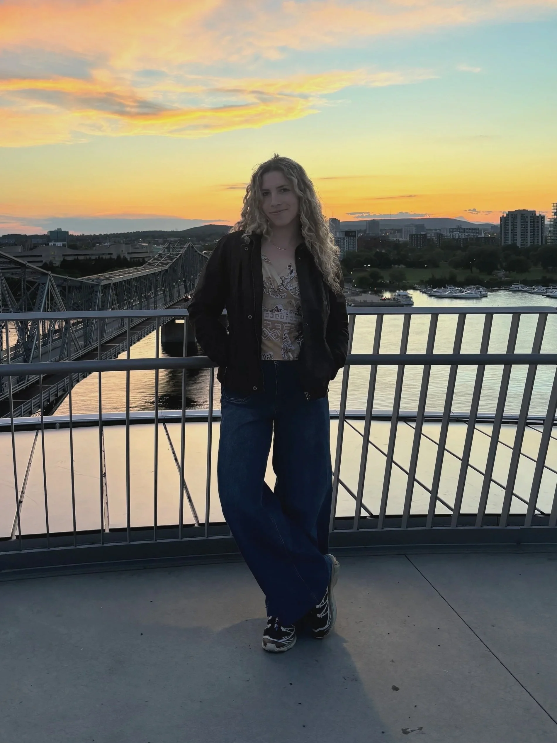 A woman with curly blonde hair stands on a bridge at sunset, with a city skyline and marina in the background.