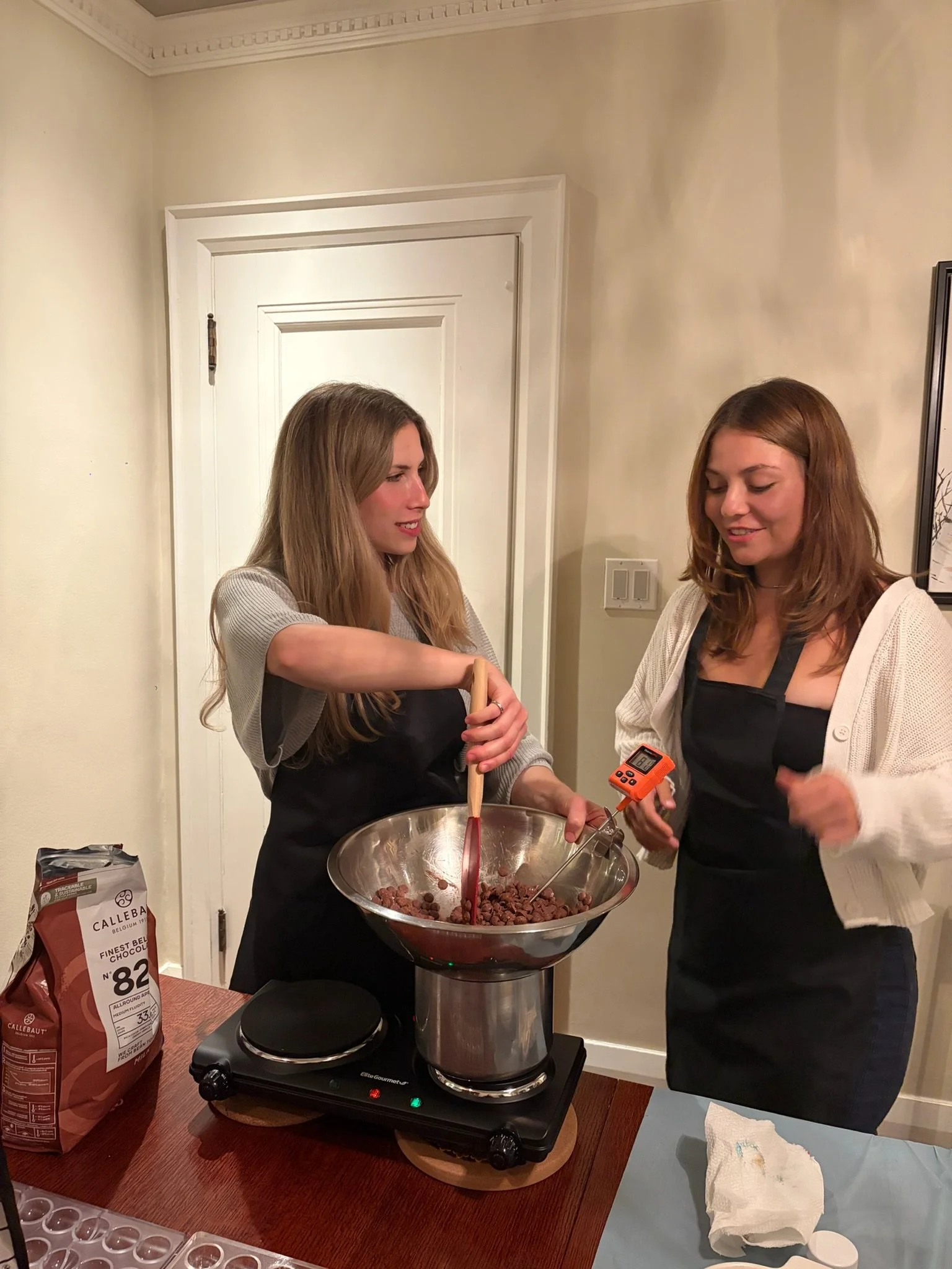 Two women are preparing chocolate in a kitchen. One woman is holding a spatula and stirring chocolate in a large mixing bowl. The other woman is holding a kitchen timer. There is a bag of Callebaut chocolate and some paper towels on the counter in front of them.