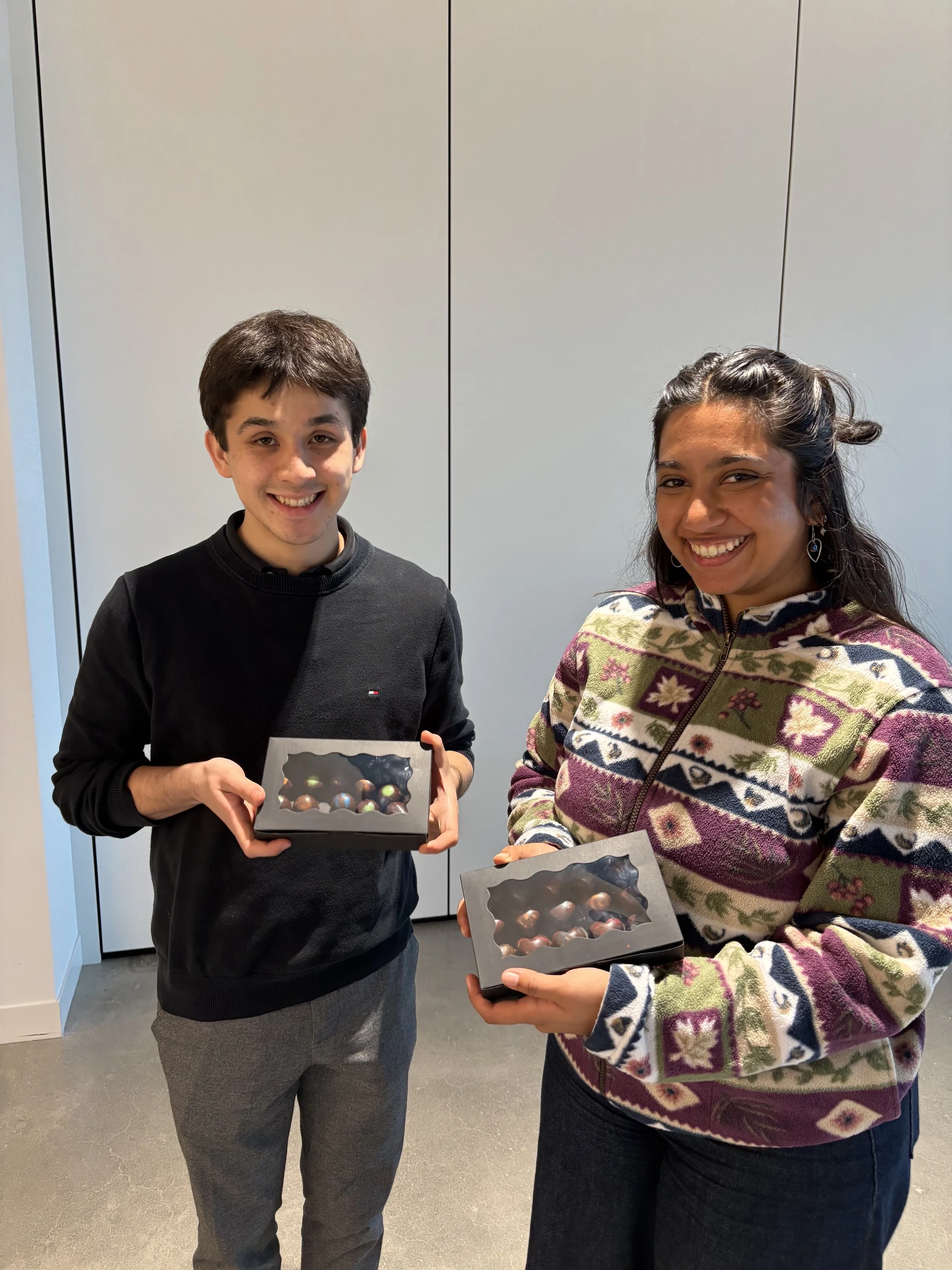 Two smiling people, one young man and one woman, standing indoors against a plain wall, each holding a box of decorated chocolates or candies.