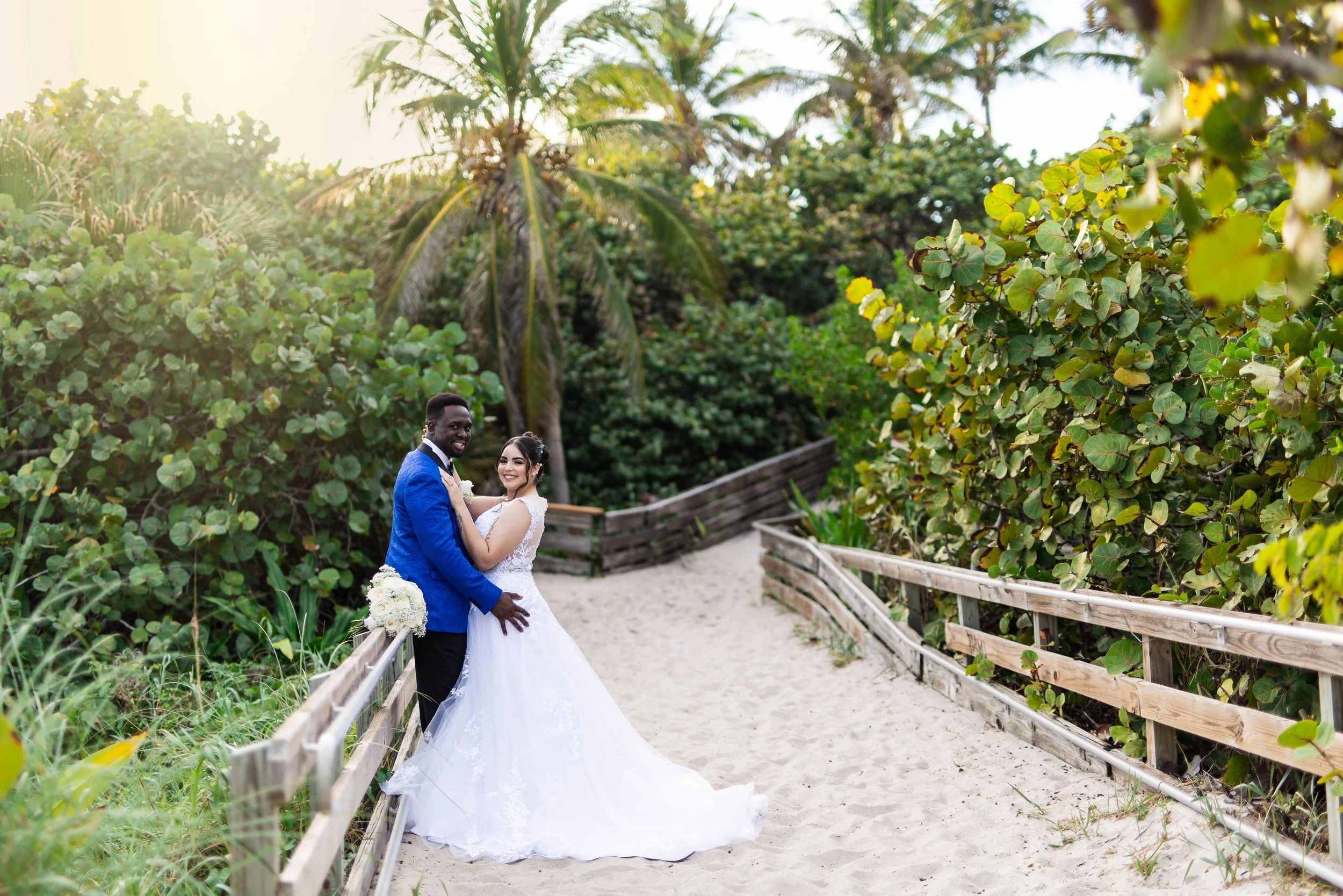 A newlywed couple in wedding attire standing on a sandy pathway surrounded by lush greenery and palm trees, smiling and embracing each other during a sunny day.