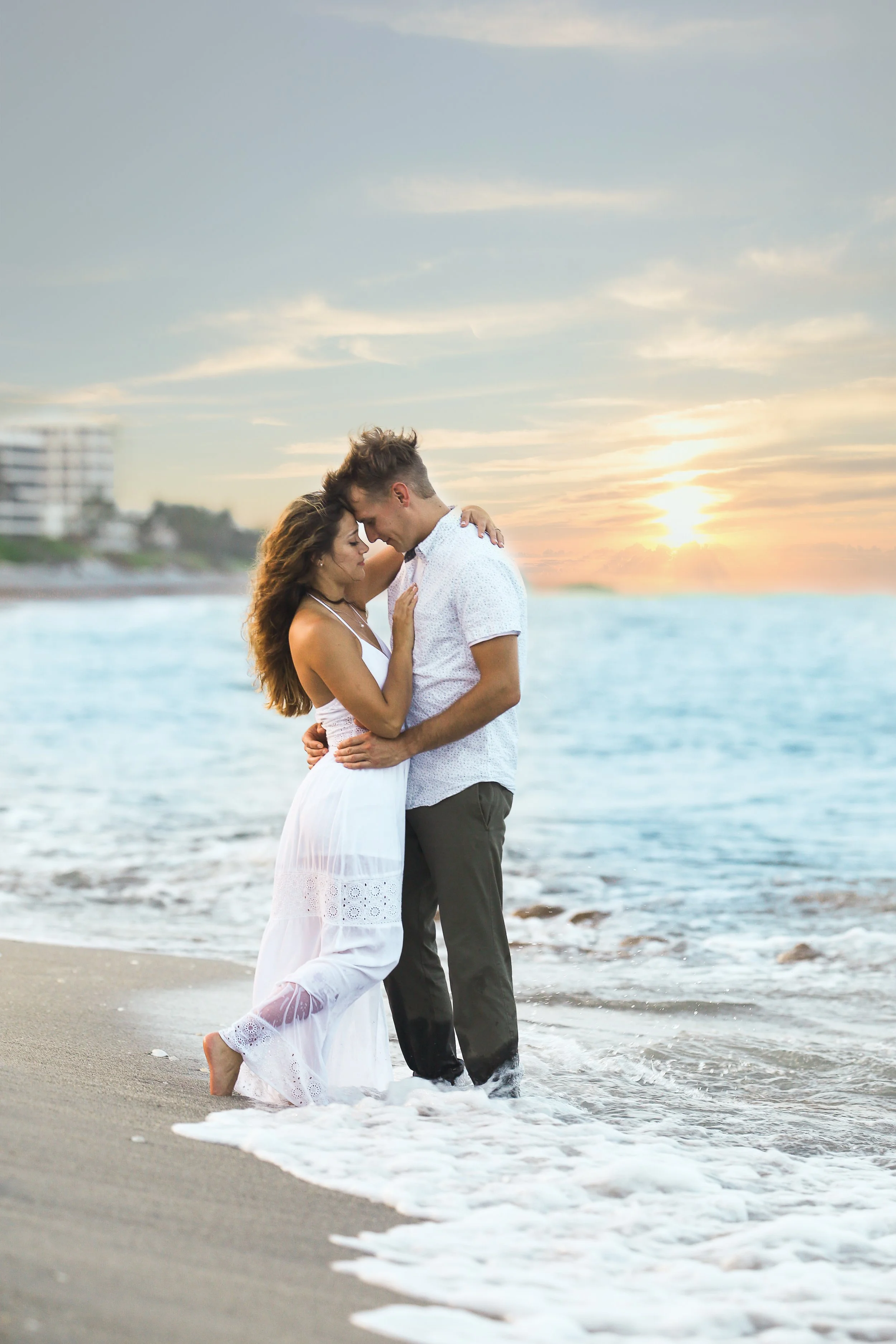 A young couple stands close together at the shoreline during sunset, with waves touching their feet. The woman wears a white dress, and the man wears a light-colored shirt and dark pants. The sky is partly cloudy with the sun near the horizon.