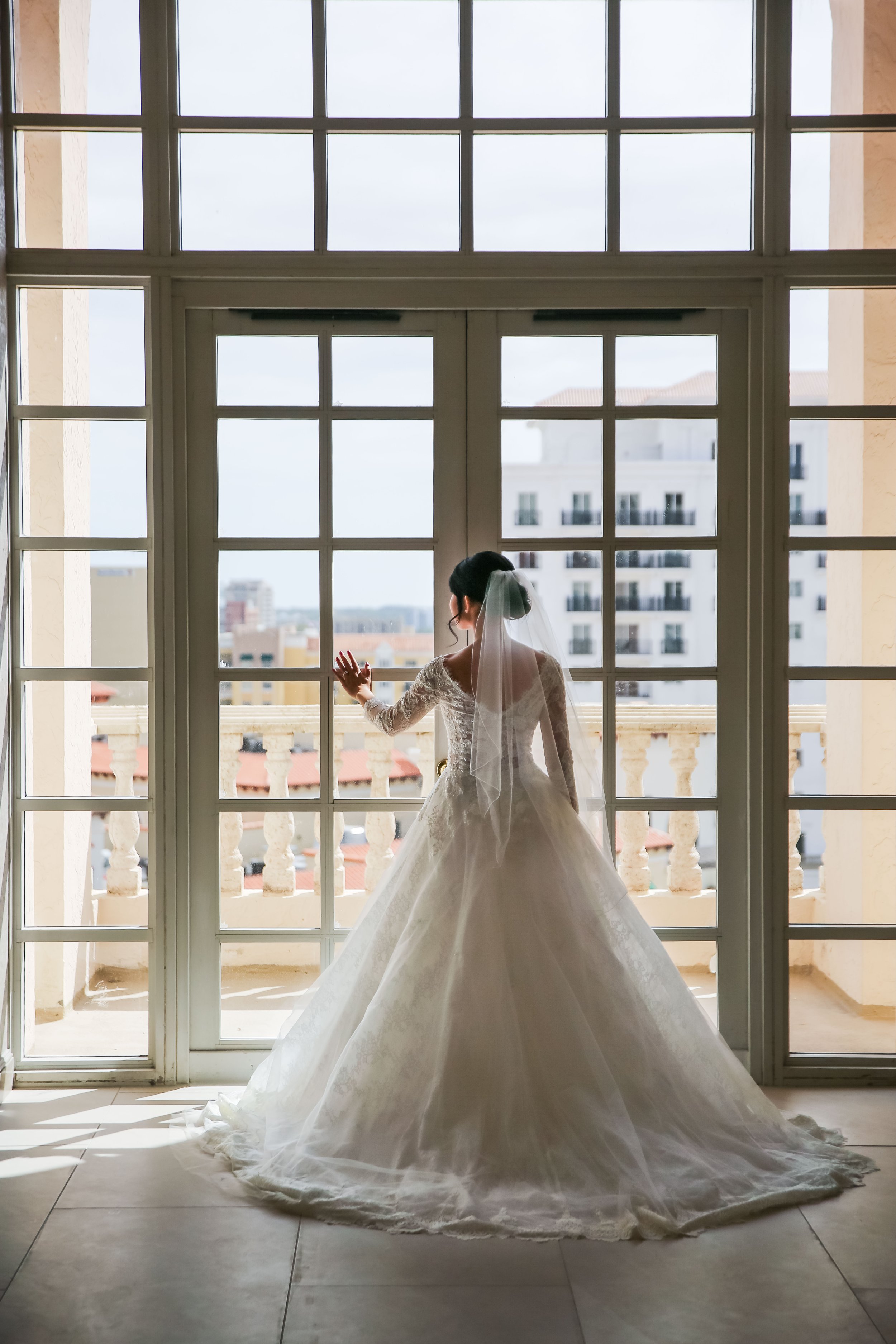 A bride in a wedding dress standing in front of large glass doors, looking out at an urban cityscape with buildings and a balcony railing visible outside.