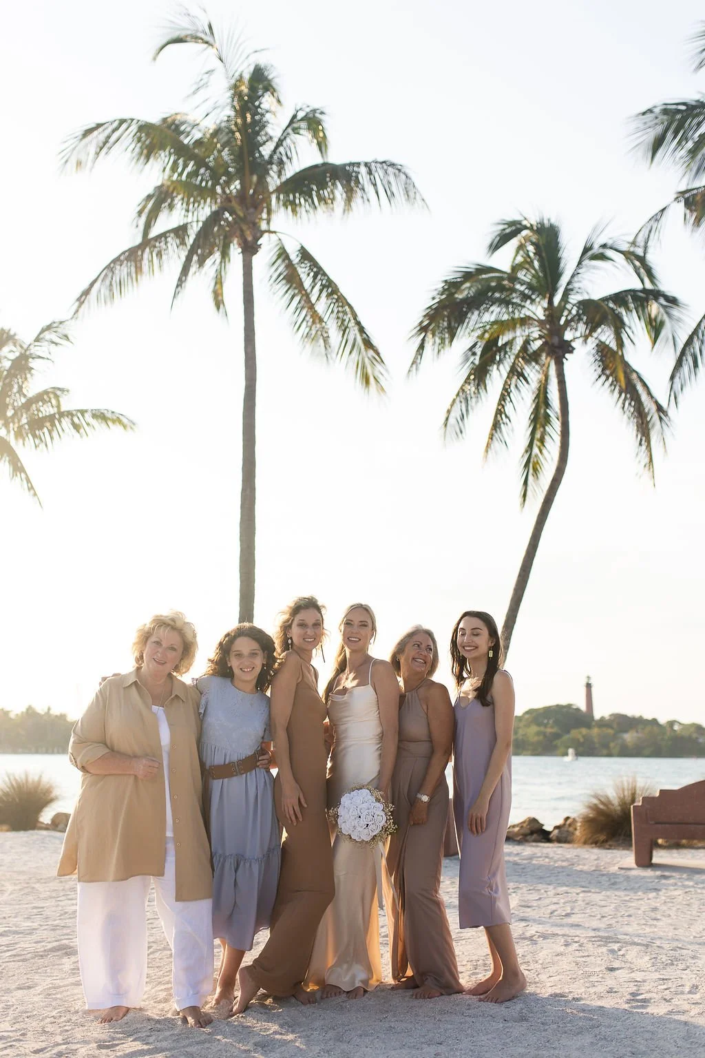 Group of six women standing on a sandy beach with palm trees, water, and a lighthouse in the background, smiling and posing together.