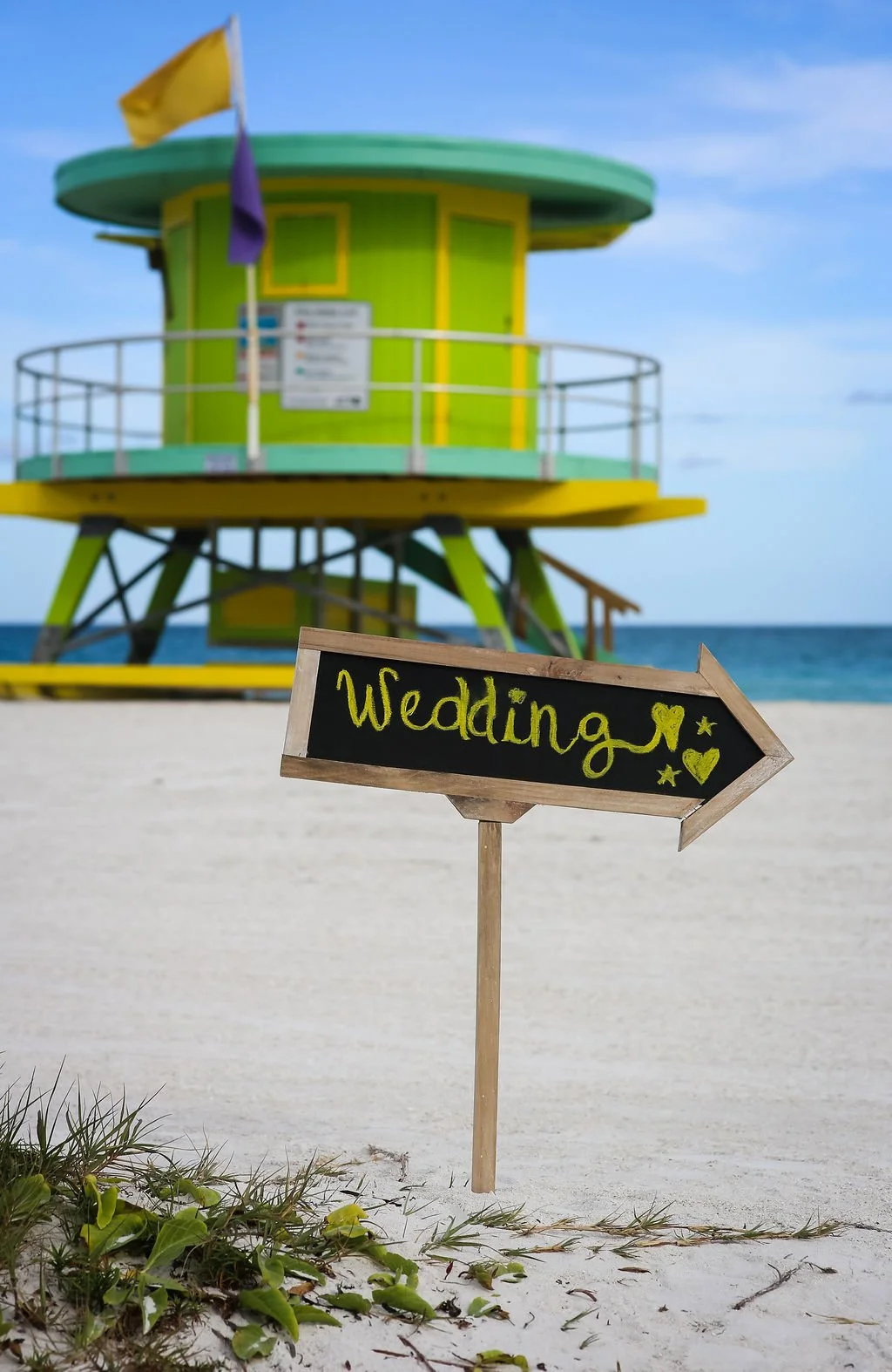 Beach scene with a green and yellow lifeguard tower and a black arrow sign on a stick with the word 'Wedding' written on it, pointing to the right.