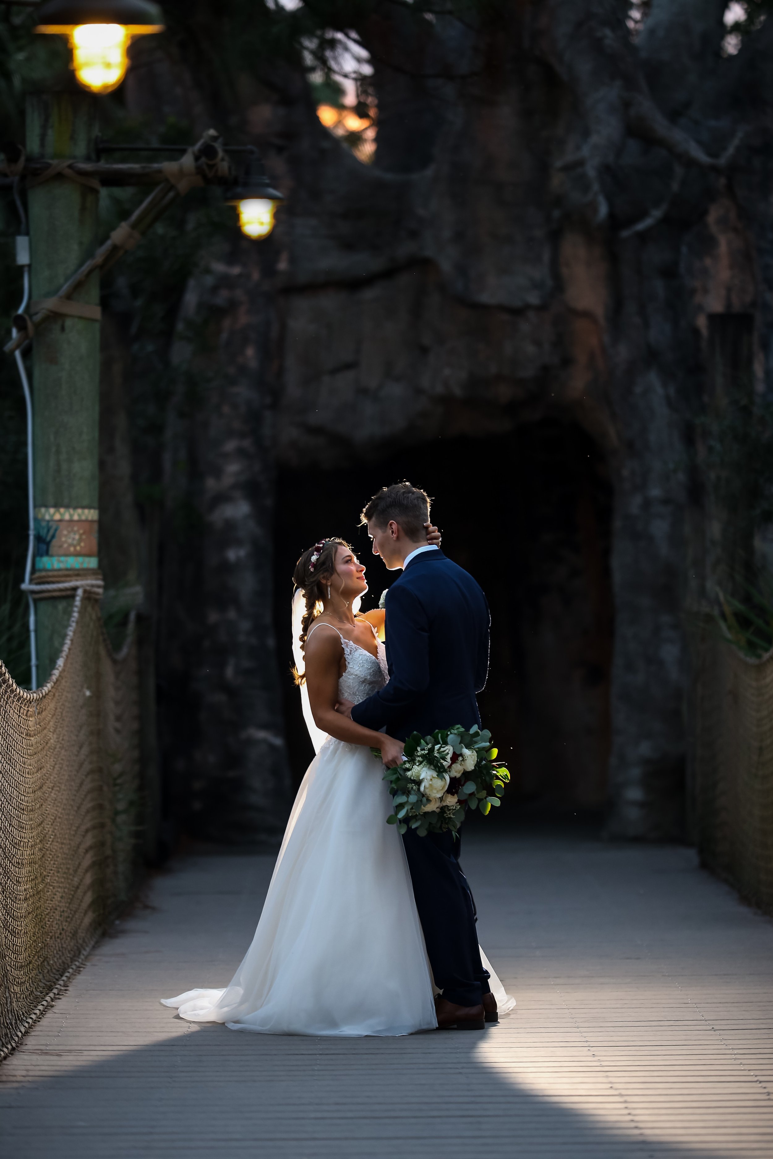 A bride and groom standing close and gazing into each other's eyes on a wooden bridge with rocky cliffs in the background, during sunset or dusk.