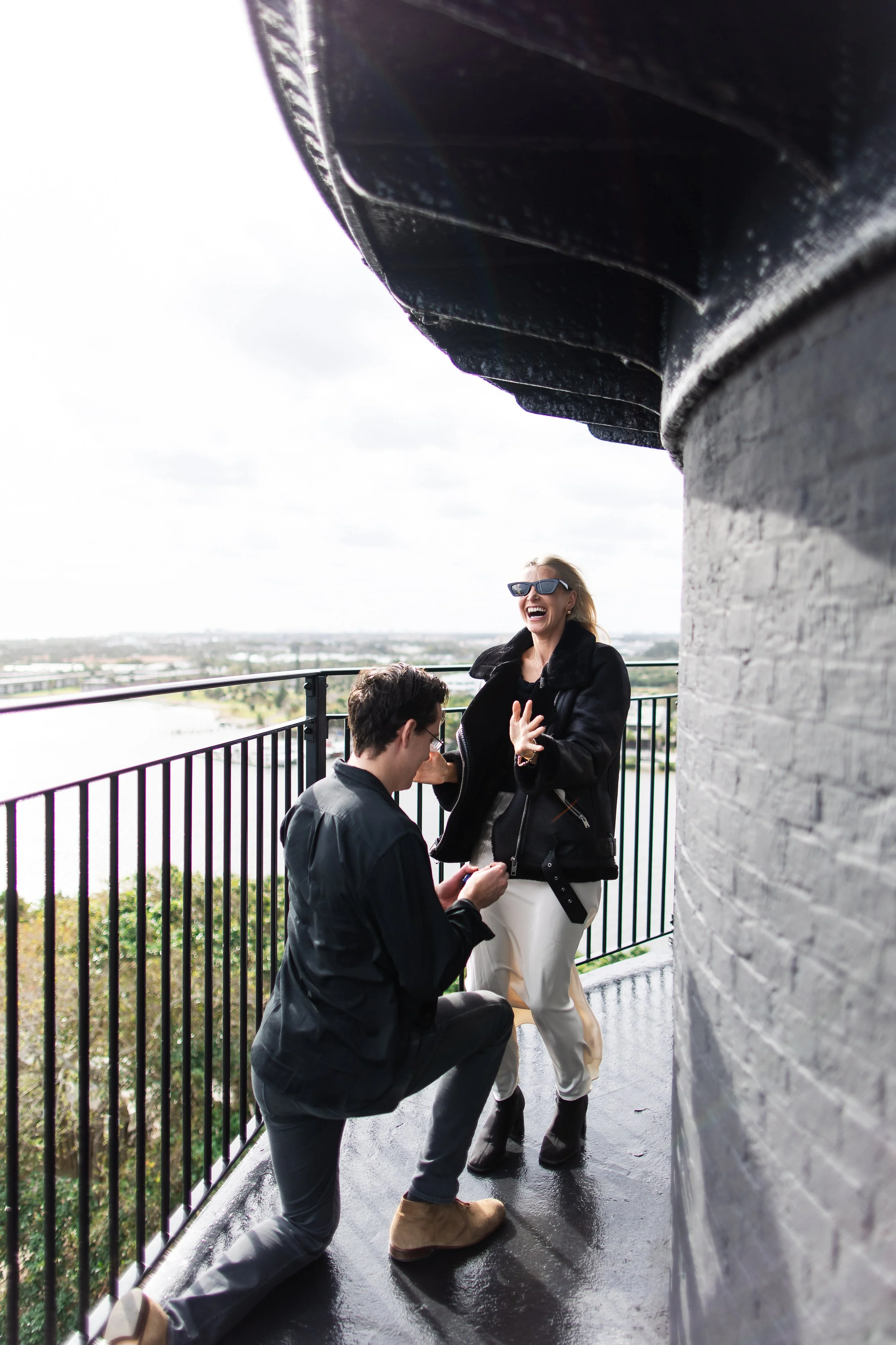 A woman laughing and talking with a man kneeling with a ring box, on a balcony with a view of a river and cityscape in the background, during daytime.