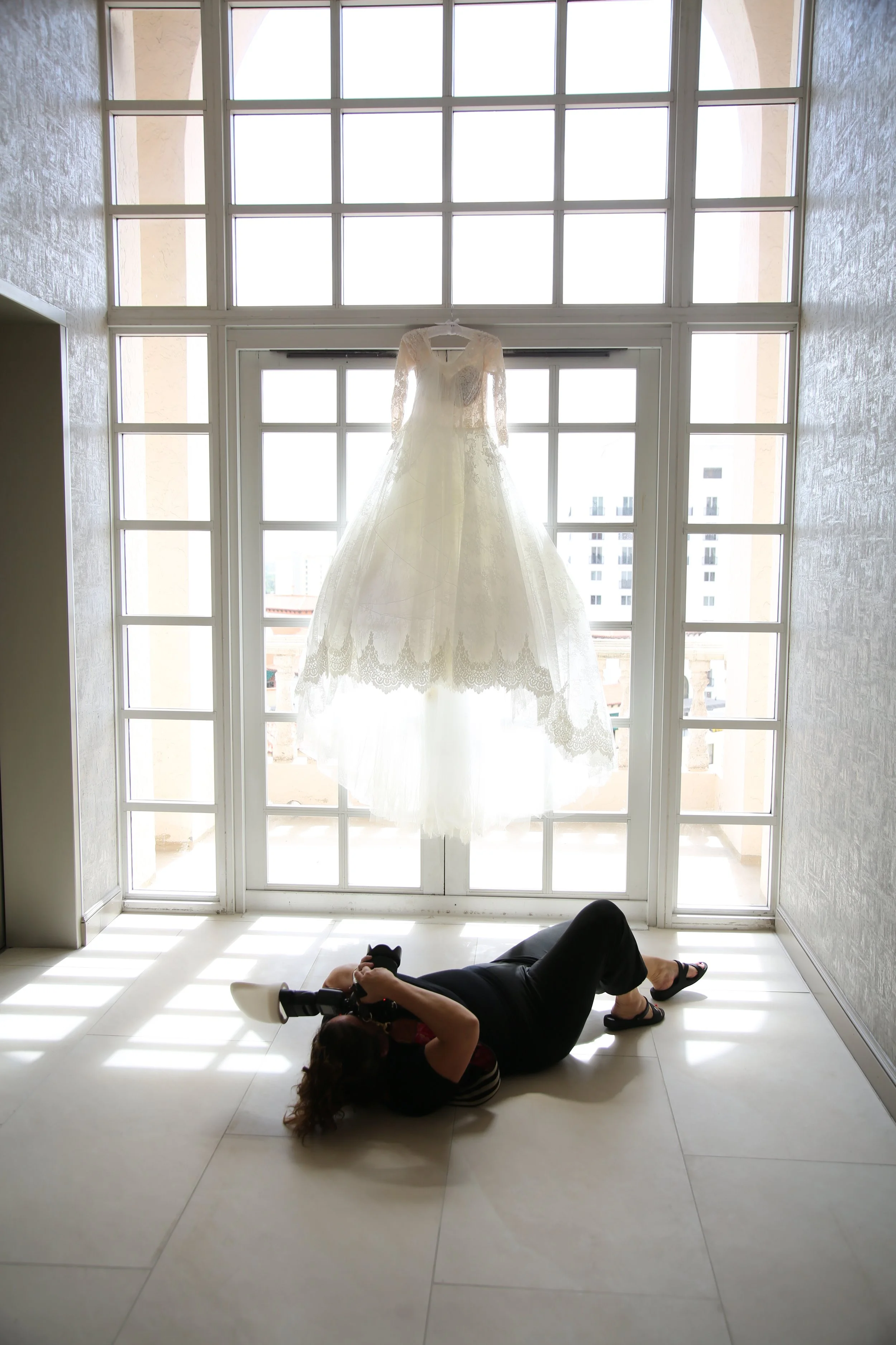 A woman lying on the floor taking a photograph of a white lace wedding dress hanging in front of a large window.