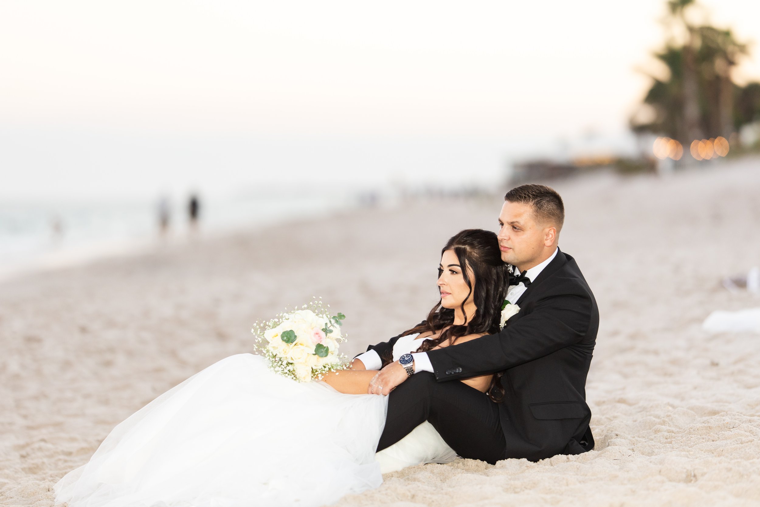 A bride and groom sitting on the beach, the groom in a tuxedo and the bride holding a bouquet of flowers, with blurred beach and palm trees in the background.