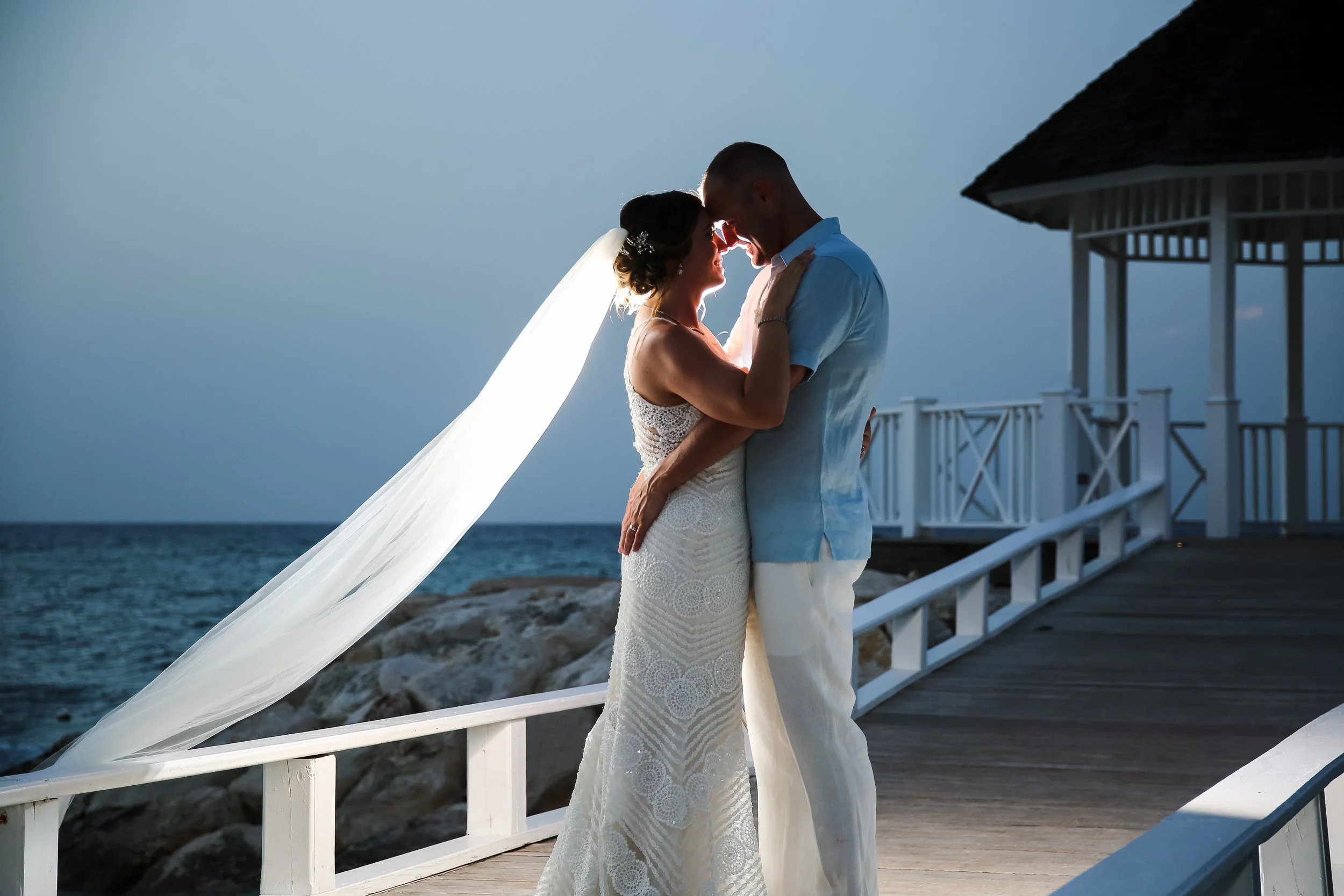 A bride and groom share an intimate moment by the beach at sunset, with the ocean in the background and a white wooden pier on the right.