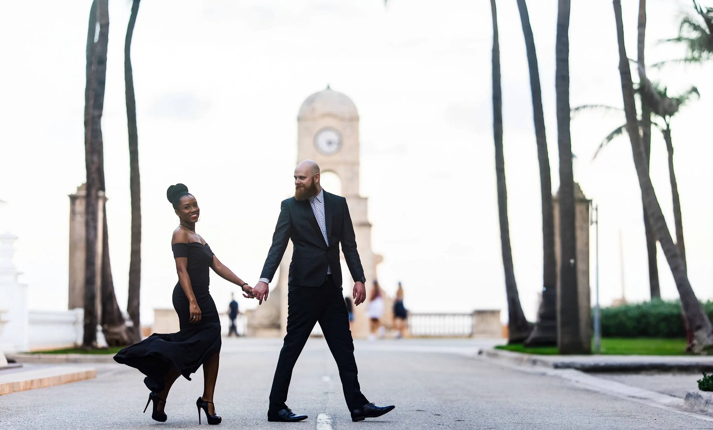 A smiling Black woman in a black off-shoulder dress and high heels holding hands with a bearded man in a suit and tie, walking together outdoors near a clock tower and tall palm trees.