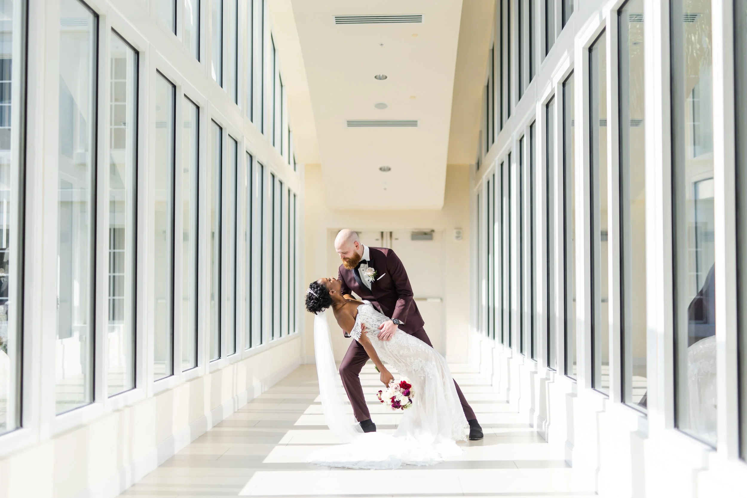 A bride and groom dancing in a modern, sunlit corridor with floor-to-ceiling windows.