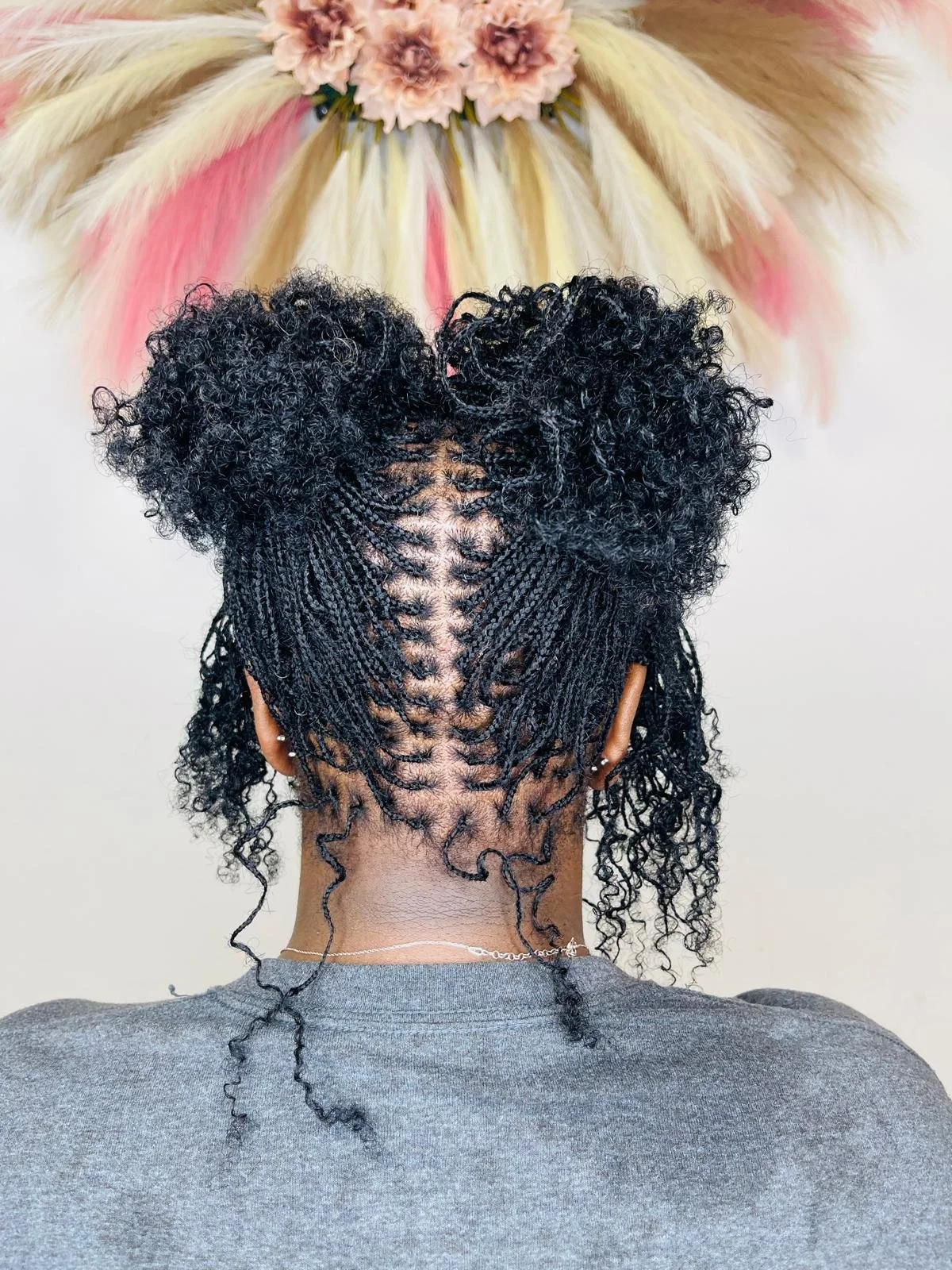 Back view of a person with small, tight braids styled into curly puffs at the top, with the rest of the hair in natural curls, and a decorative wall hanging with pink flowers and multicolored pampas grass in the background.