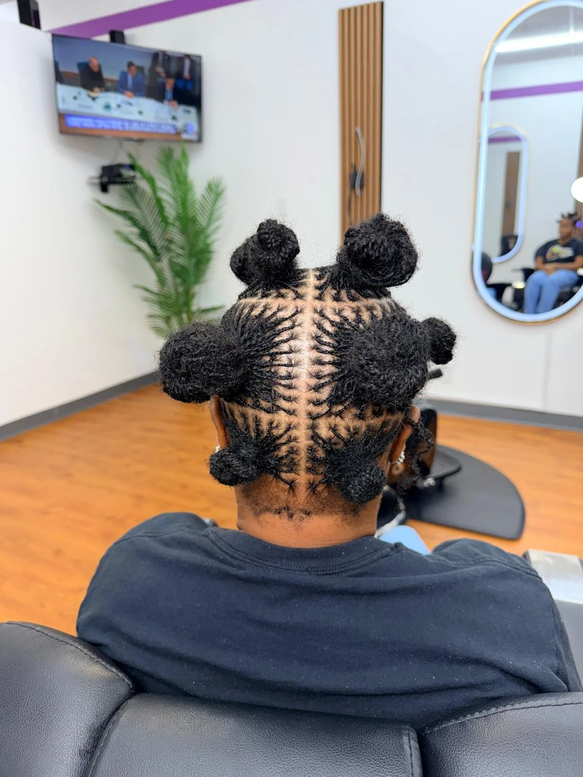 Person with a unique braided hairstyle featuring multiple large bun sections arranged on their head, sitting in a salon chair.
