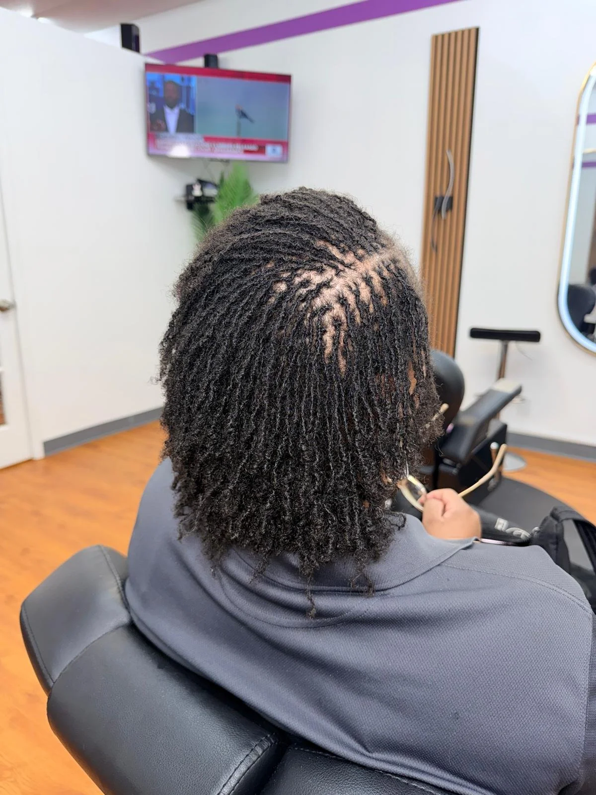 Rear view of a woman with natural, textured hair sitting in a salon chair, wearing a gray hoodie, in a modern hair salon. In the background, a television is mounted on the wall and a mirror is visible.