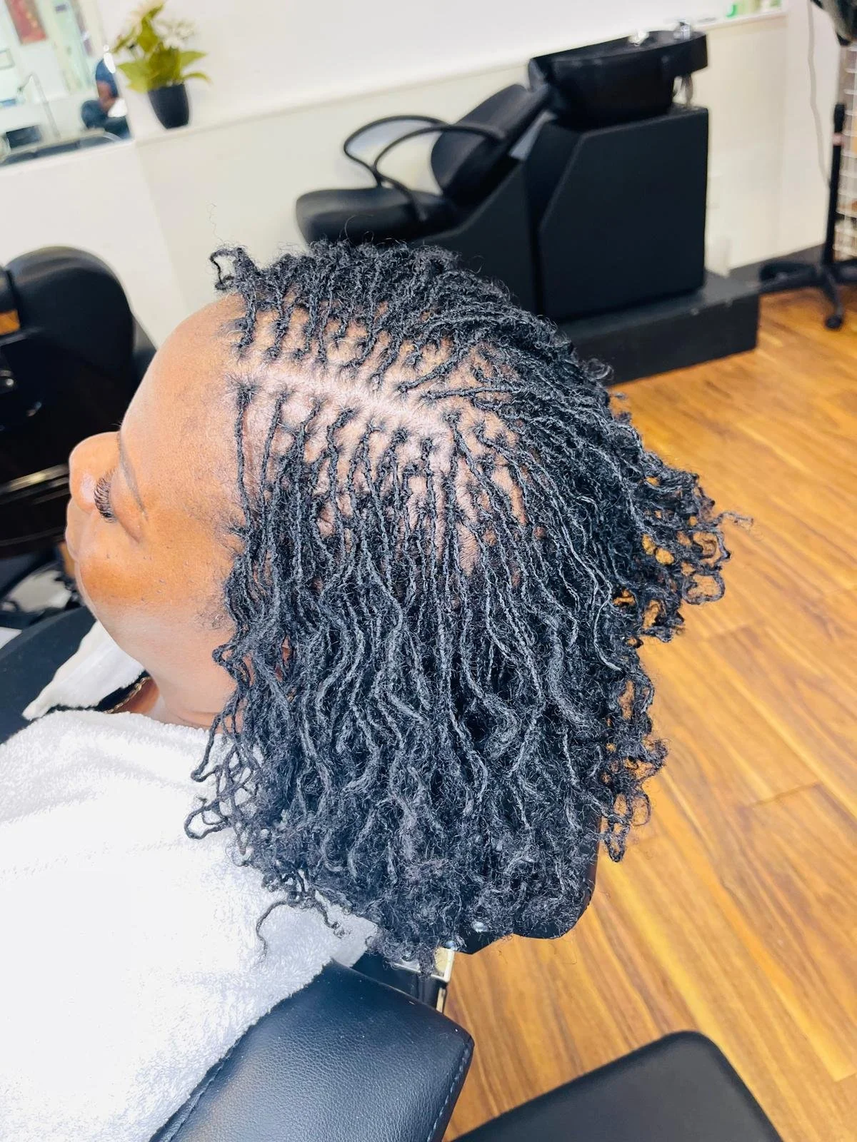 A woman with short, natural textured hair sitting in a salon chair. Her hair is styled in twist-out curls. The salon has hardwood floors and black salon equipment in the background.