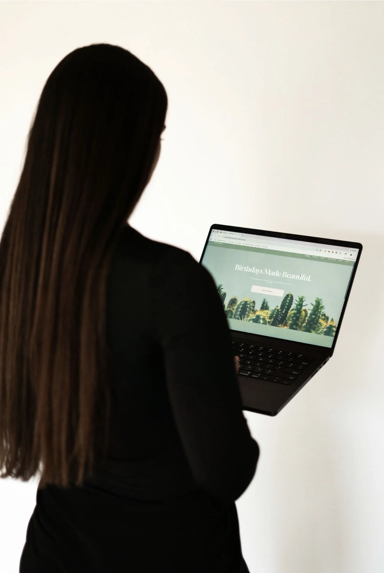 A woman with long hair, dressed in black, is looking at a laptop screen displaying a website with a green background and text that reads 'Birthdays Made Beautiful' and a picture of cacti.