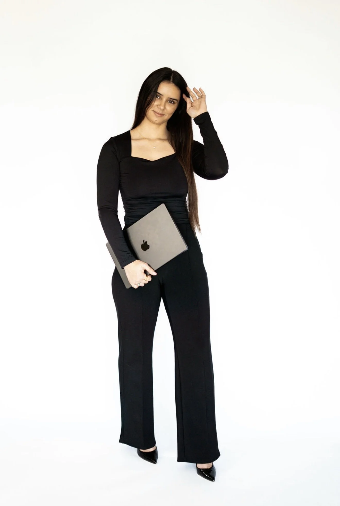A woman in black professional attire holding a silver MacBook laptop, standing against a white background.