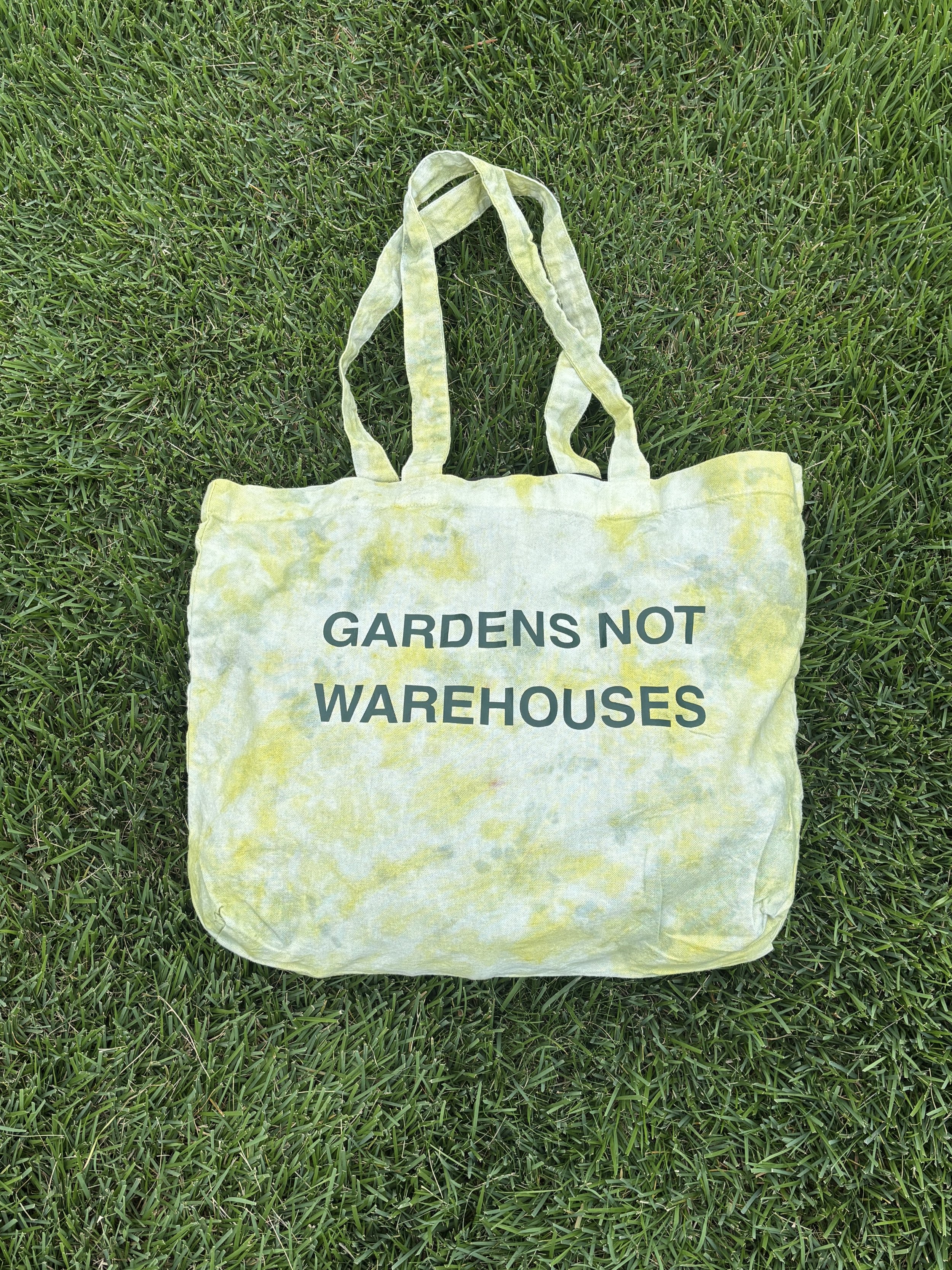 A yellow fabric tote bag with the words "GARDENS NOT WAREHOUSES" printed on it, lying on green grass.