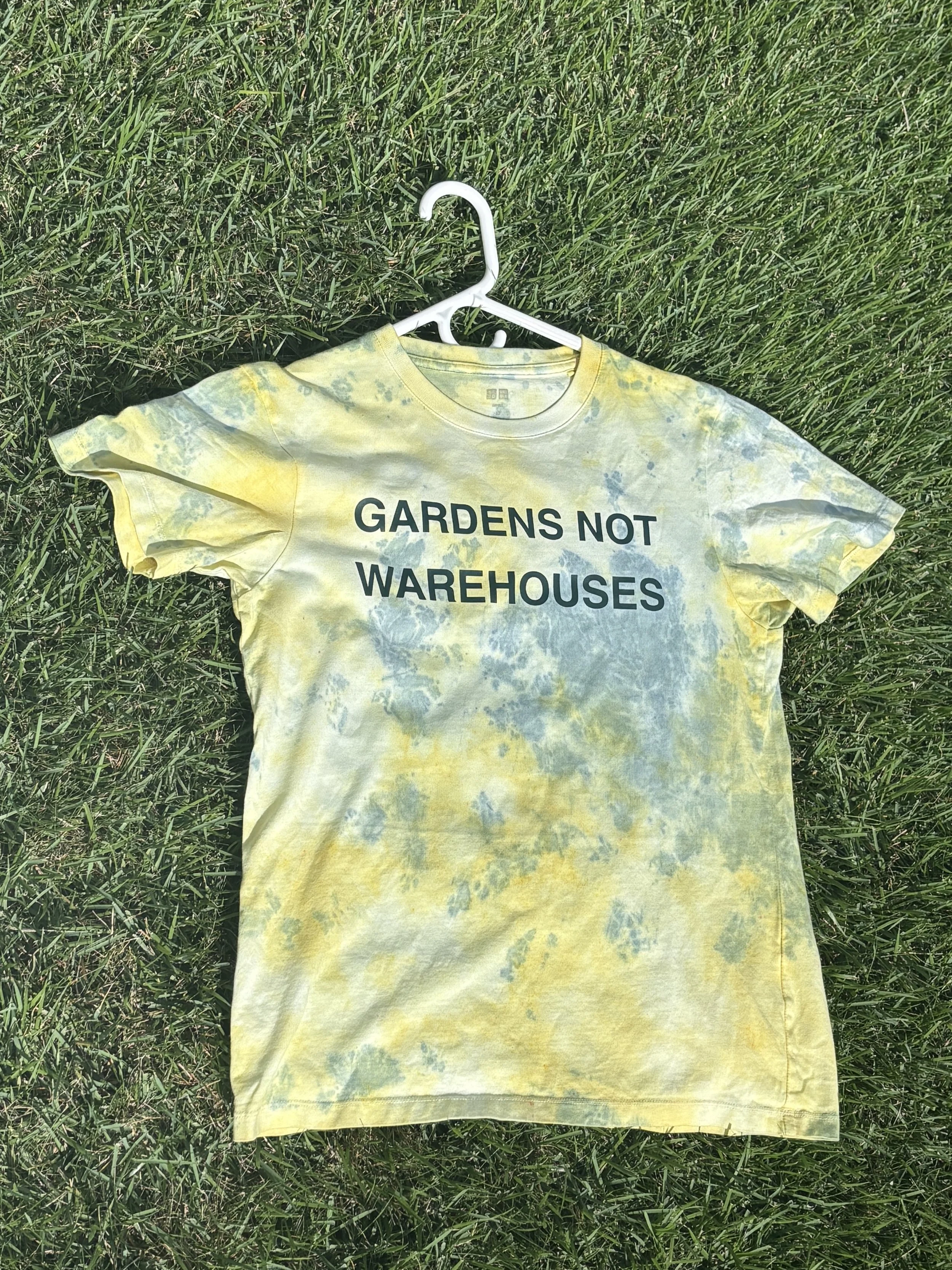 Yellow tie-dye T-shirt with black text that reads "GARDENS NOT WAREHOUSES," laid on green grass and hung on a white hanger.