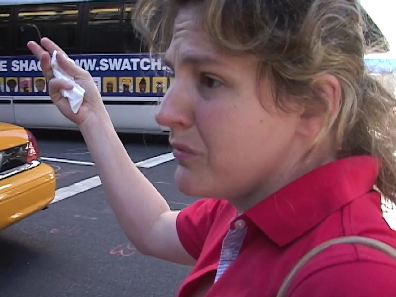 A woman with curly blonde hair in a red shirt holding a tissue, standing near taxis and a billboard that reads 'www.swatch' with images of watch faces.