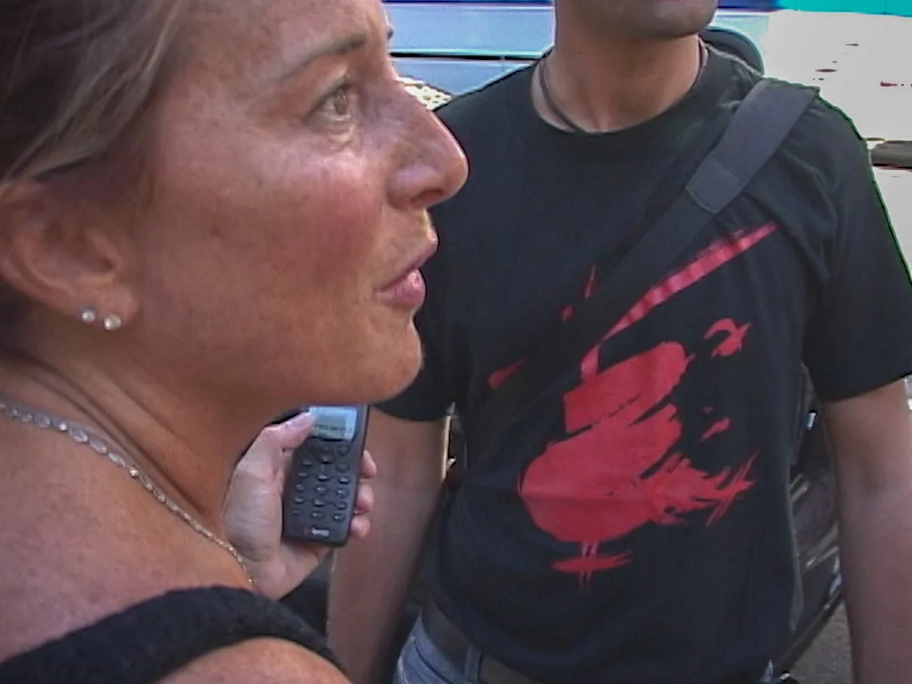 Close-up of a woman with blonde hair and earrings, holding a remote control, standing next to a person wearing a black t-shirt with a red graphic design and a strap across their chest.