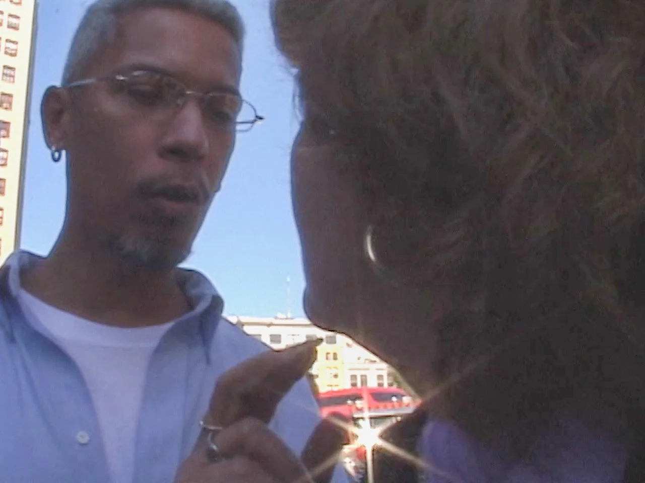 A man wearing glasses and a white shirt appears to be in a heated conversation with a woman beneath a clear blue sky, with city buildings and a red vehicle visible in the background.