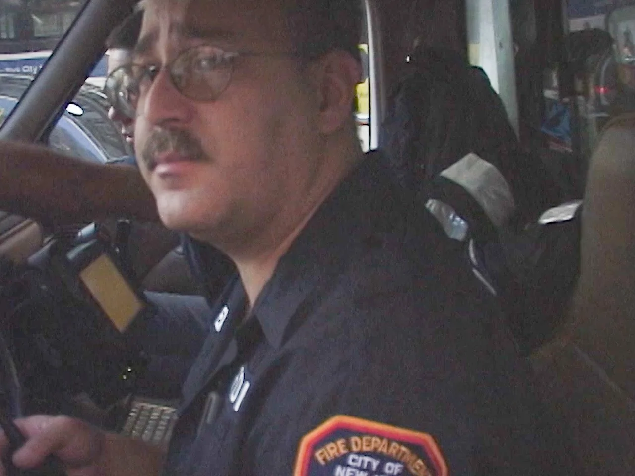 A firefighter in uniform with a badge that reads 'Fire Department City of New York' is sitting inside a vehicle, looking at the camera, holding a device, with emergency vehicles and equipment visible in the background.