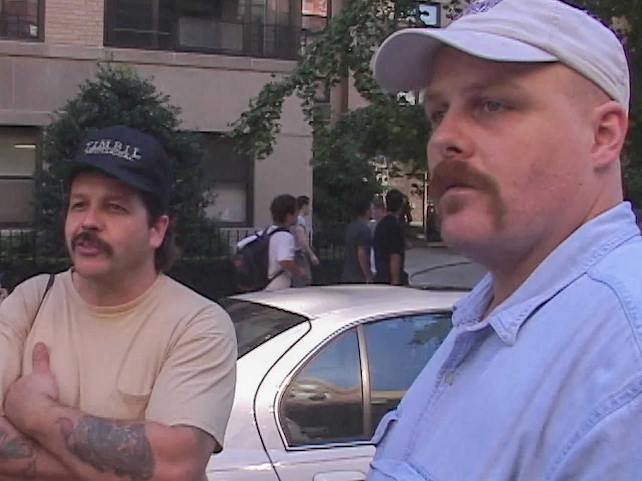 Two men with mustaches and hats standing outside near a car and a group of people walking on the sidewalk in an urban area.