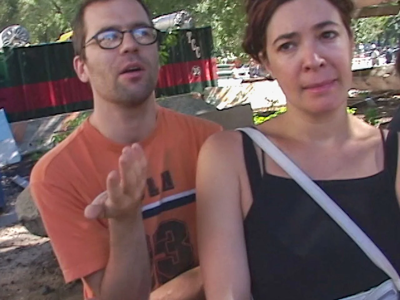 A man with glasses and an orange t-shirt is speaking and gesturing with his right hand. A woman with short brown hair, wearing a black top and a white strap, is standing next to him, looking slightly away. They are outdoors with trees and a fence in 