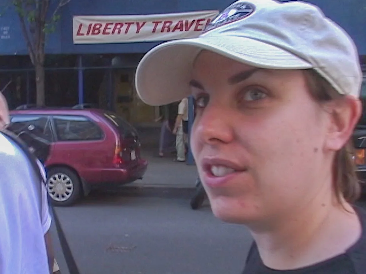 Close-up of a woman with a beige baseball cap, outside on a city street with a reddish-maroon station wagon and a banner reading 'Liberty Travel' in the background.