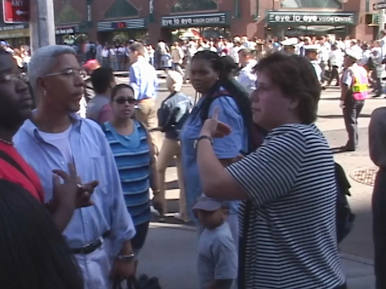 A group of people standing and talking on a busy city street during the daytime, with many more pedestrians and storefronts in the background.