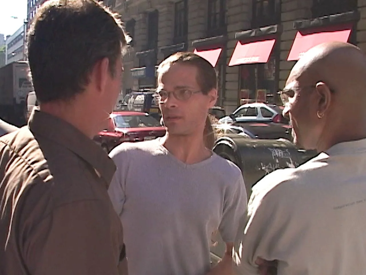 Three people standing on a city sidewalk engaged in conversation, with buildings and parked cars in the background.