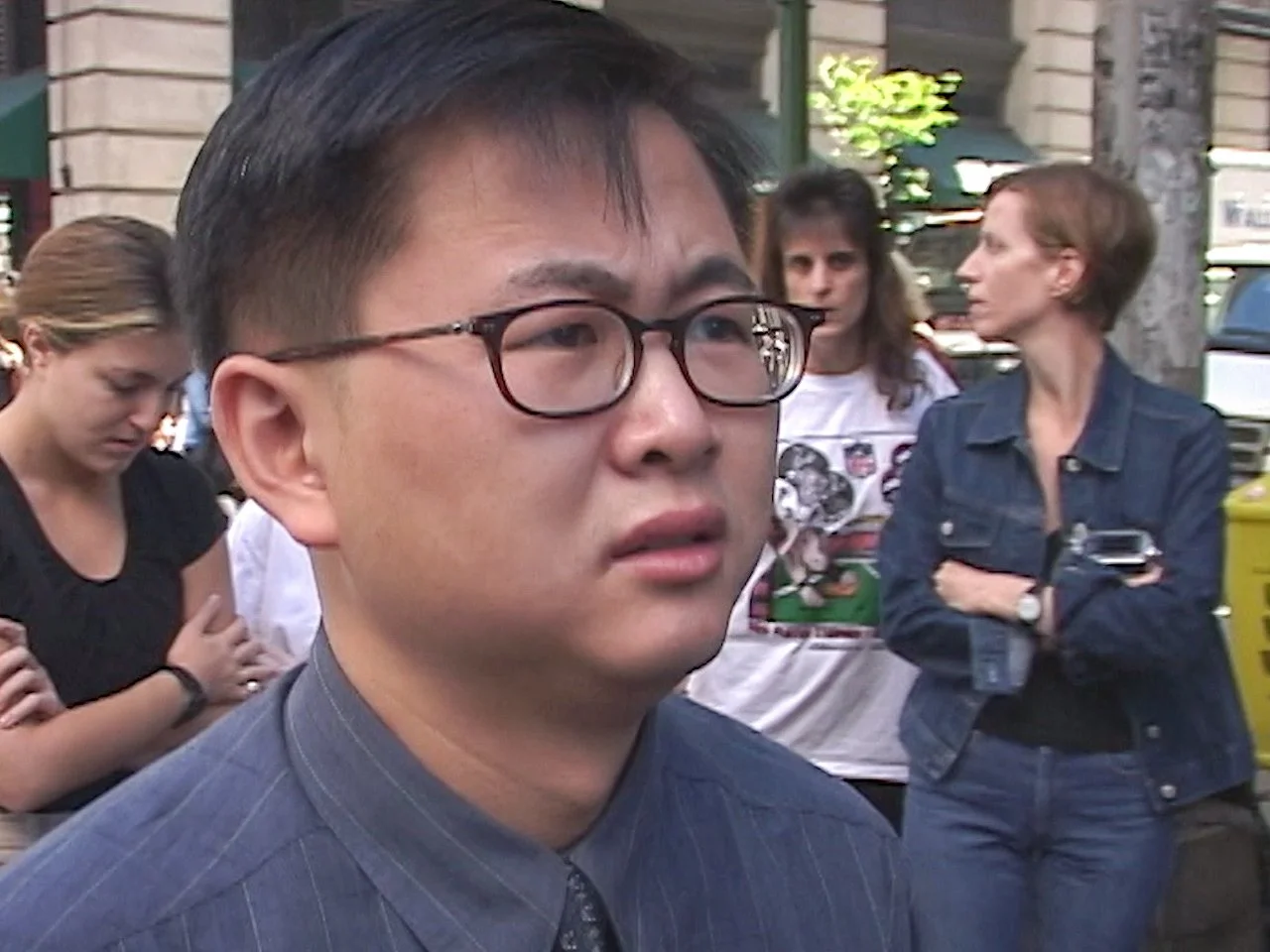A young Asian man with glasses, wearing a gray pinstripe shirt, appears to be outdoors on a city street. In the background, several women are seen, including one with a white T-shirt with a graphic and two with denim jackets, standing among trees and