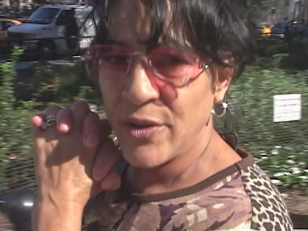 A woman wearing pink sunglasses and leopard print top stands outdoors, touching her cheek with her hand, with a parking lot and green bushes in the background.