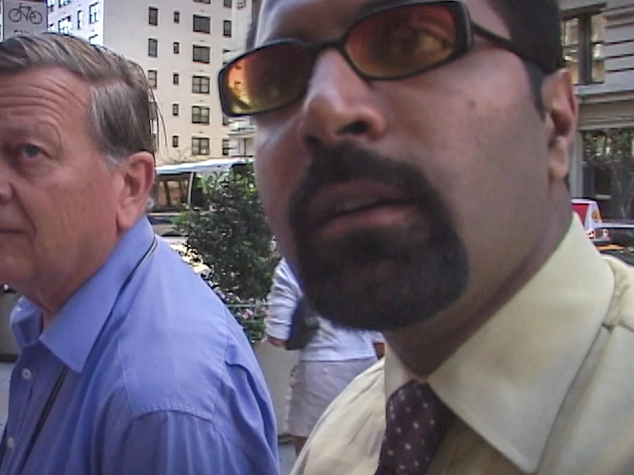 Close-up of two men outdoors in an urban setting, one wearing sunglasses and a yellow shirt, and the other with gray hair in a blue shirt, with buildings and other people in the background.