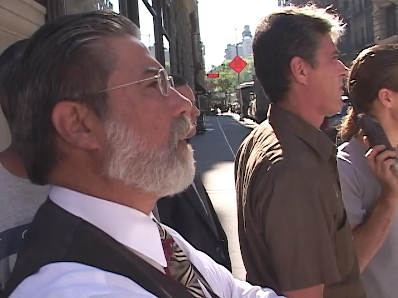 Three people standing side by side on a city sidewalk on a sunny day. The man in the foreground has glasses, a white shirt, and a beard. The man in the background is talking on a cell phone. The woman is partially visible.