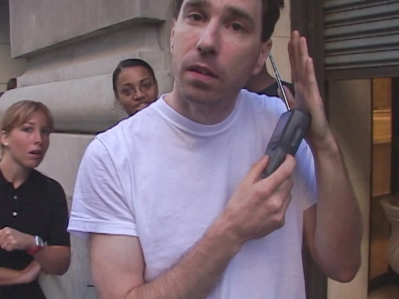 A man in a white t-shirt using a walkie-talkie or radio device, with two women standing behind him looking surprised or confused, outside near a building with stone and metal surfaces.