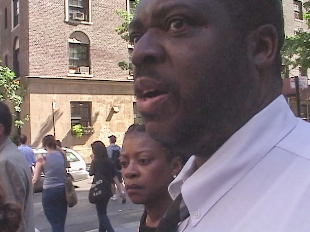 Close-up of a man in a white shirt talking outdoors, with people walking on the street and buildings in the background.