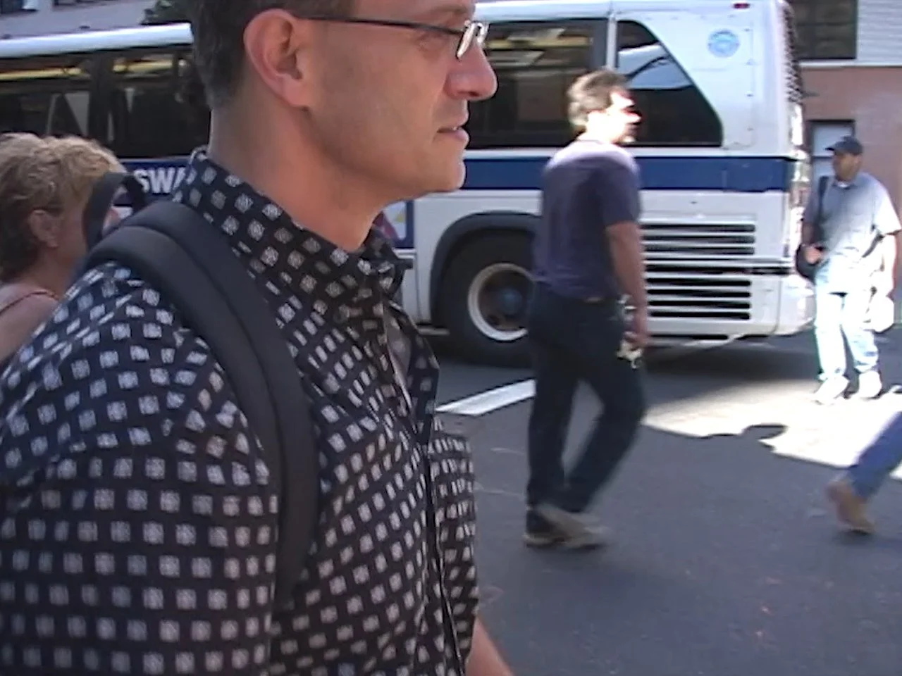A man with glasses and a patterned shirt carrying a backpack walking on a busy city street with a bus in the background.