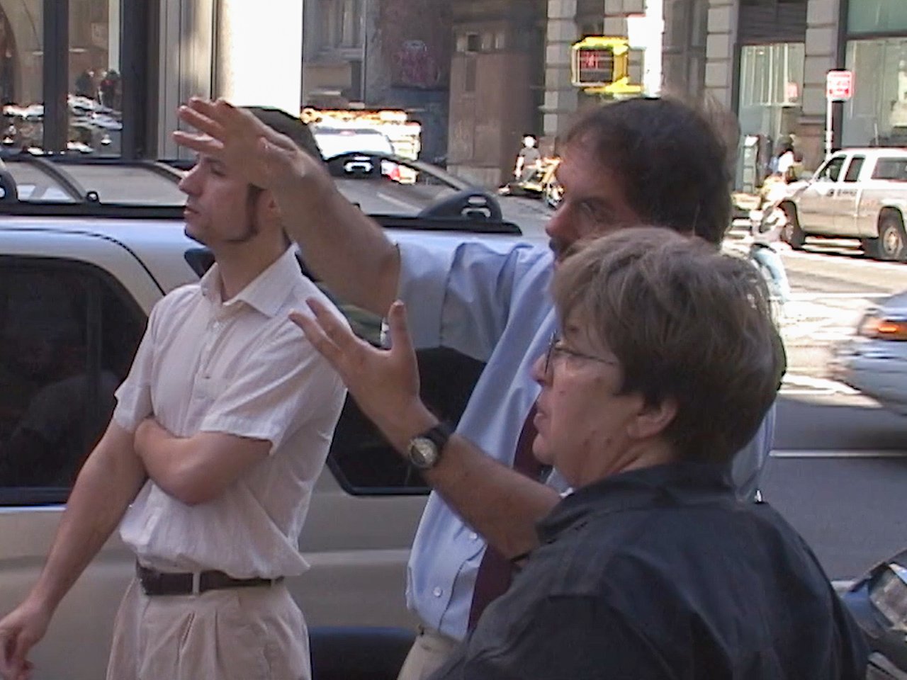 Three people appear to be having a discussion or debate on a city street, with cars and buildings in the background.
