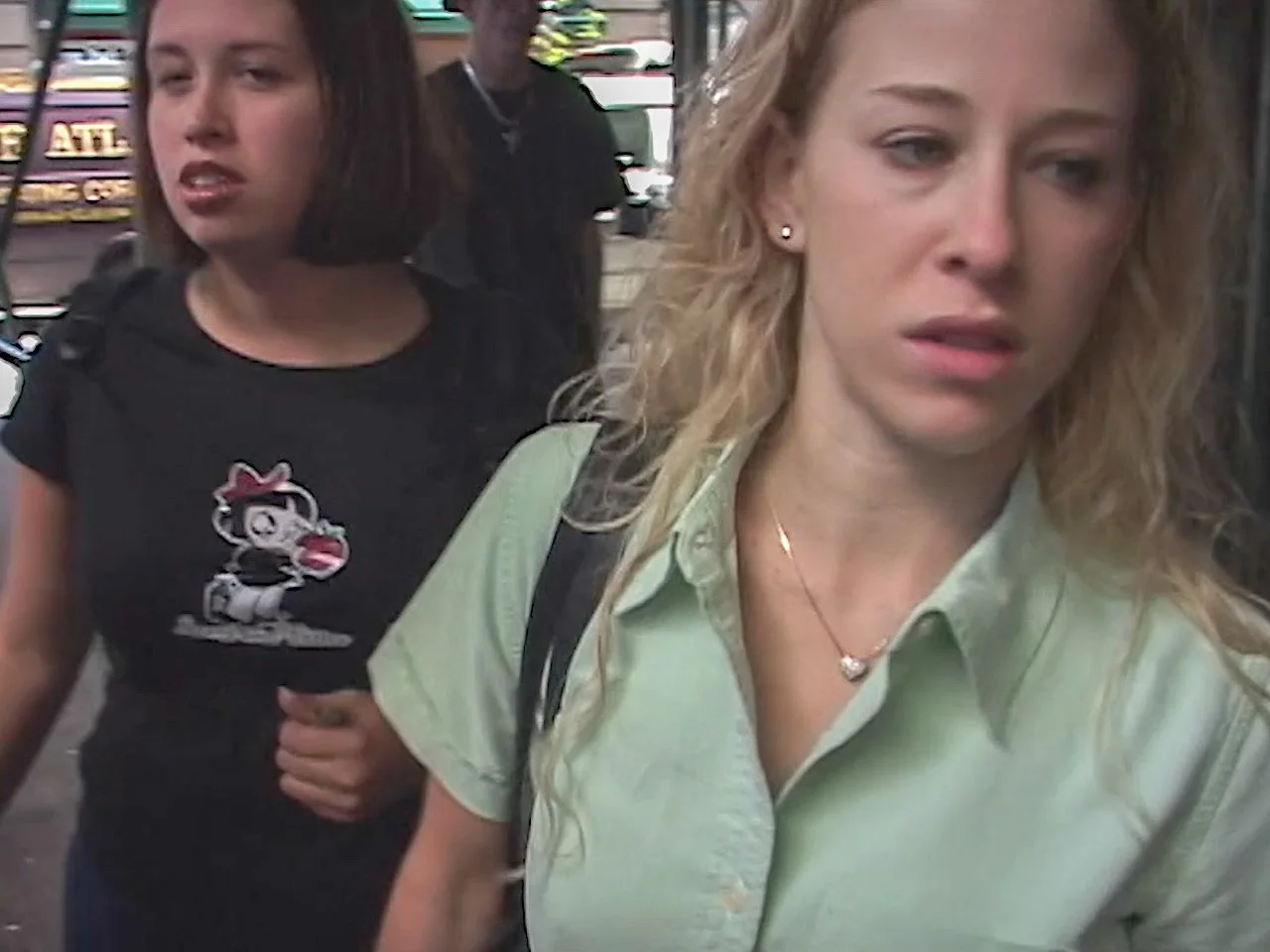 A woman with curly blonde hair and wearing a light green shirt is in the foreground, looking to her right with a concerned expression. A young girl with straight brown hair, wearing a black T-shirt with a cartoon character, is walking behind her on t